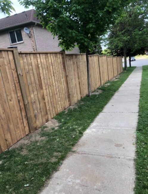 A residential street view with a tall wooden fence, sidewalk, grass, and trees, beside a brick house on a sunny day.