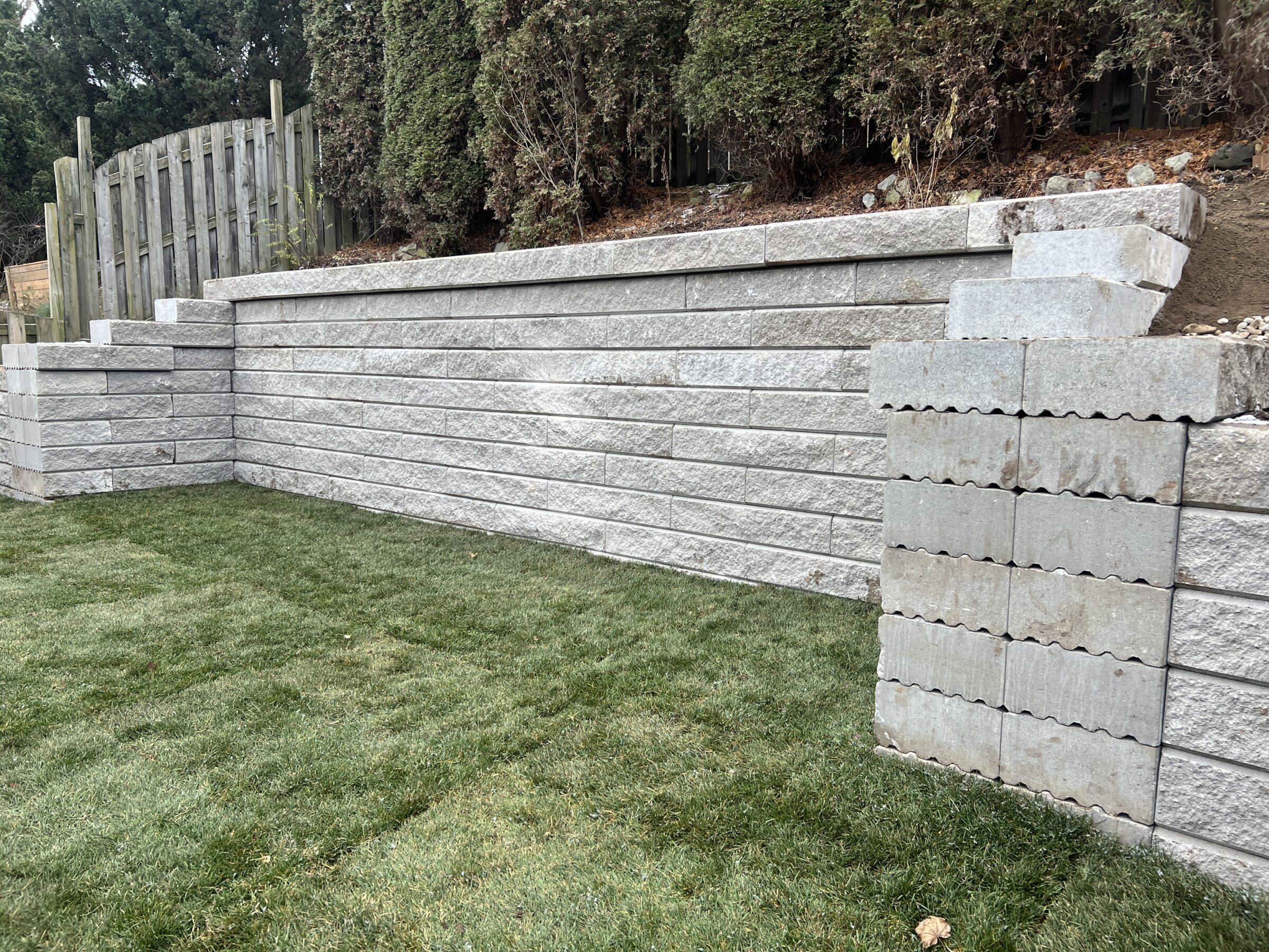 A stone retaining wall with neat, horizontal blocks stands beside a lush, green lawn and a wooden fence, bordered by dense shrubs.