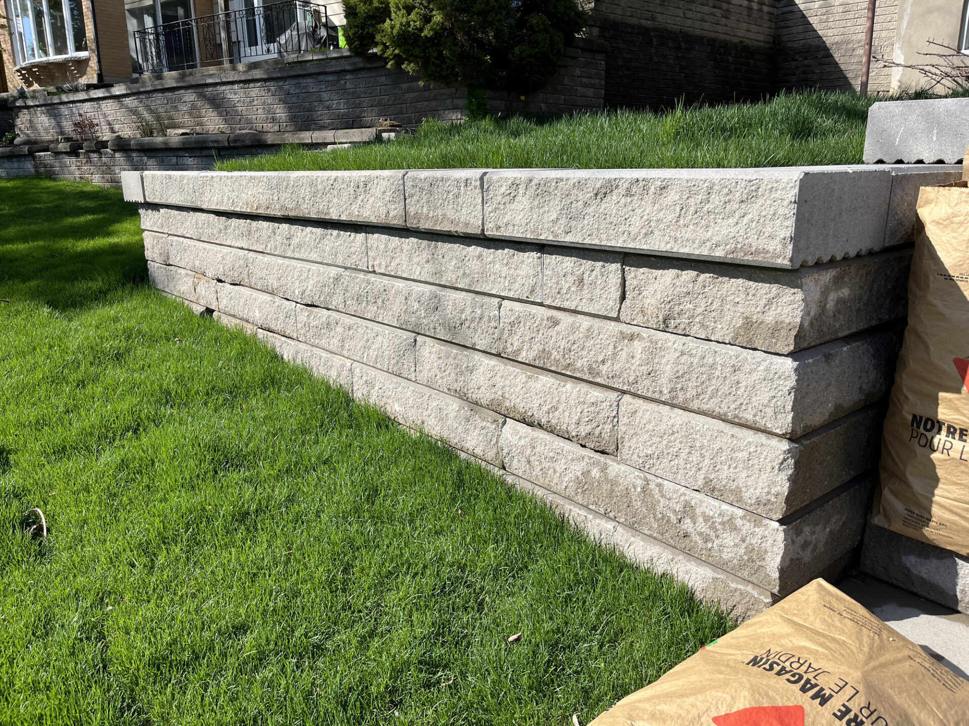 A stone retaining wall beside a grassy lawn. Bags of gardening material are nearby, with brick steps visible leading to a house entrance.