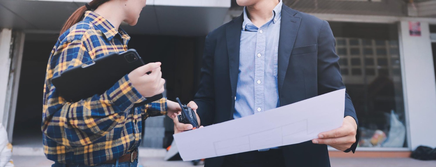 Two persons are outside, discussing plans. One holds blueprints; the other has a tablet and keys. They appear engaged in conversation.