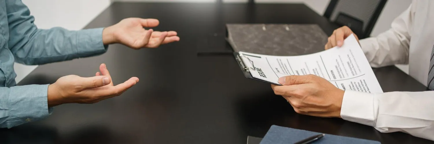 Two people sitting at a table in a meeting; one person holds a resume, and there's a discussion taking place.