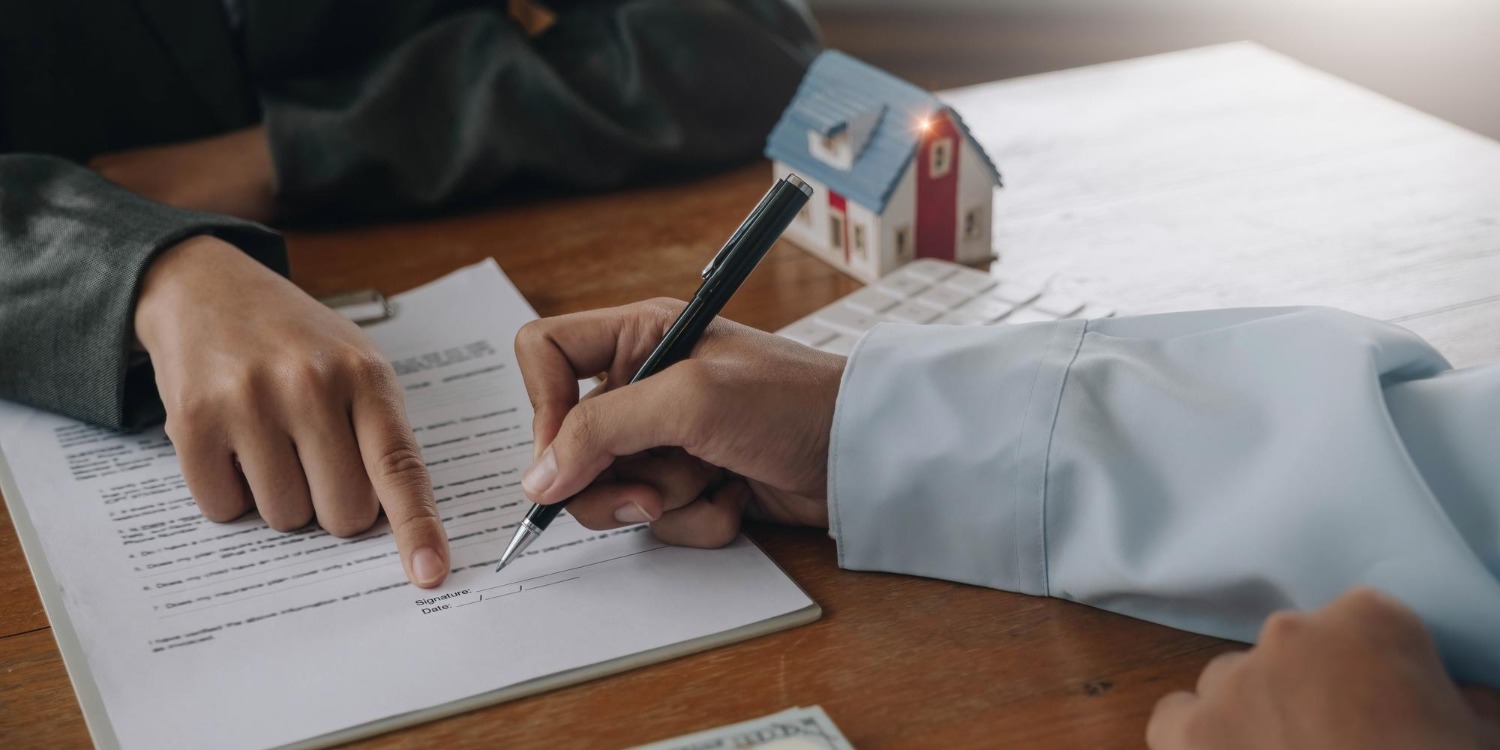 Two people review and sign a contract on a wooden table, near a small model house, suggesting a real estate transaction.
