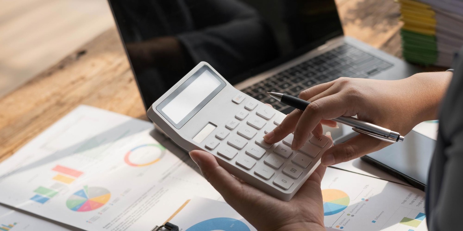A person uses a calculator at a wooden desk with charts, documents, and an open laptop, focusing on financial analysis and data management.