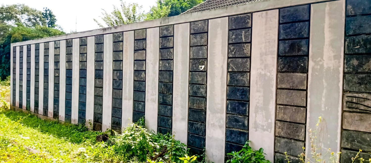 Long concrete wall with vertical black and white sections, surrounded by lush greenery in a sunny outdoor setting. No recognizable landmarks.