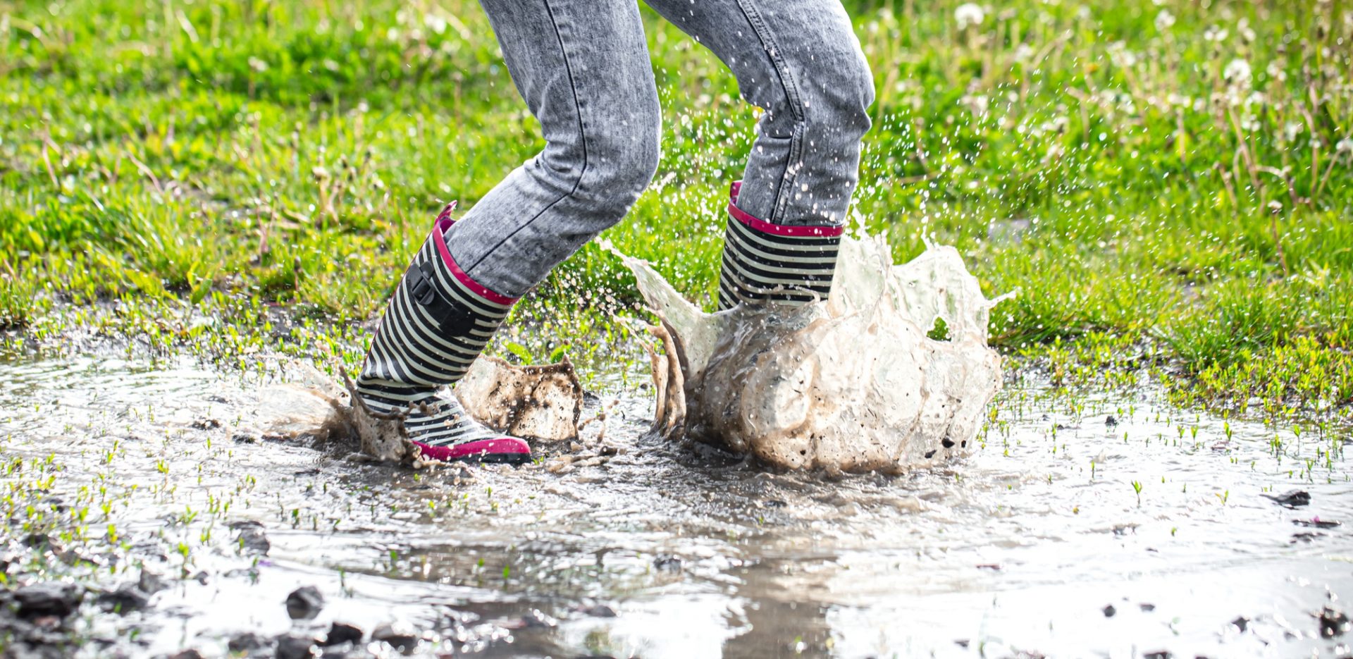 A person splashes in a muddy puddle, with water droplets and stripes of colorful rain boots visible in a grassy area.